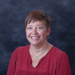A white woman with short brown hair and blue eyes wearing a dark red v-neck shirt and butterfly necklace