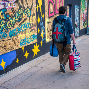 Person walking down city sidewalk with several bags and graffiti in the background