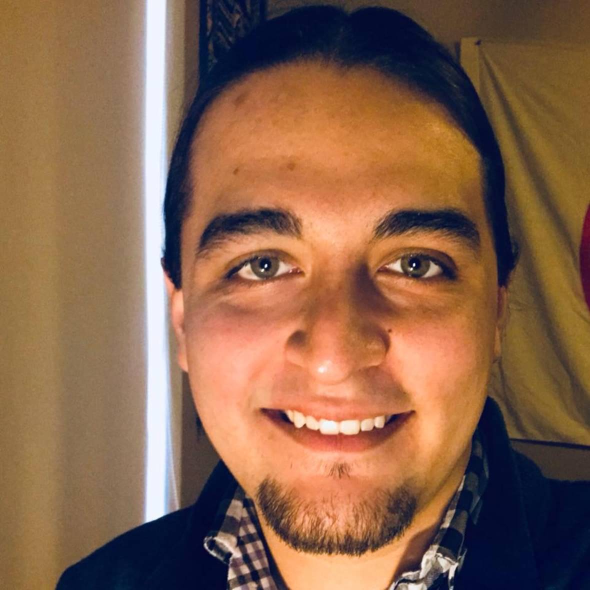 A close-up of a smiling young man with green eyes, tied-back long dark hair, and a goatee