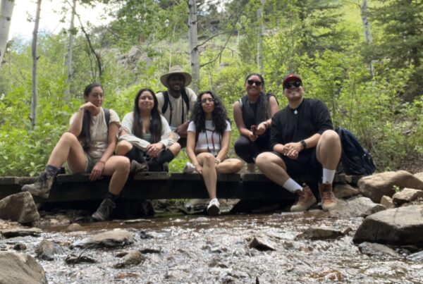 Six people of color in hiking gear sit next to a creek