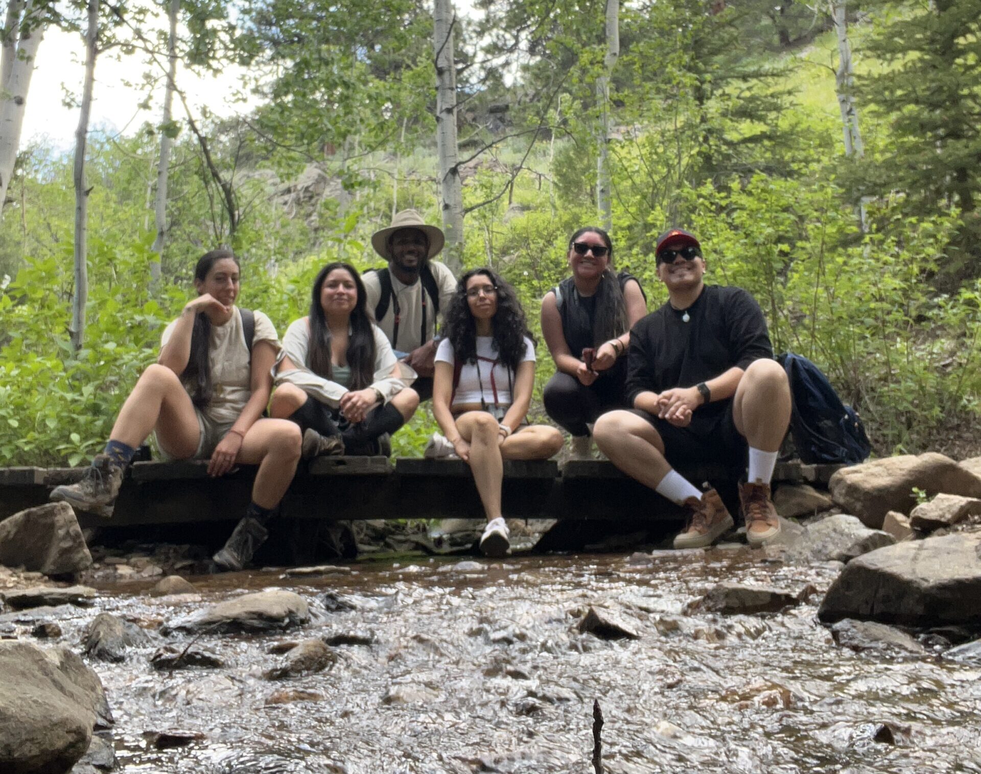 Six people of color in hiking gear sit next to a creek