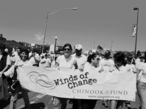 Group of people march down a street in a large crowd holding a banner that says Winds of Change Chinook Fund