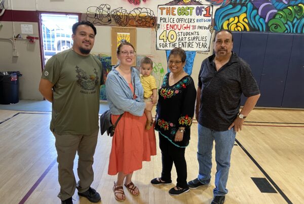 Four people stand in school gym with poster on the wall calling for less funding for police riot gear and more funding for school arts