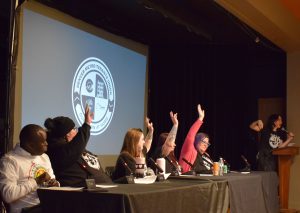 Five people sit behind a table on stage, four of them have one hand raised