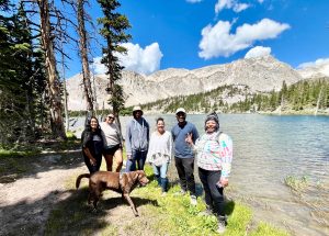 Six people of color and a brown dog stand next to a river with trees, hills, and blue sky in the background.