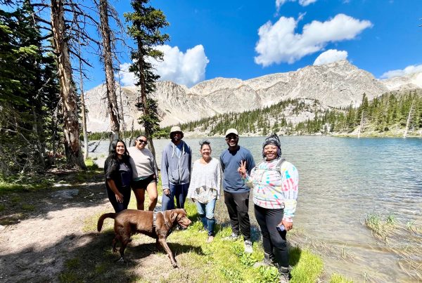 Six people of color and a brown dog stand next to a river with trees, hills, and blue sky in the background.